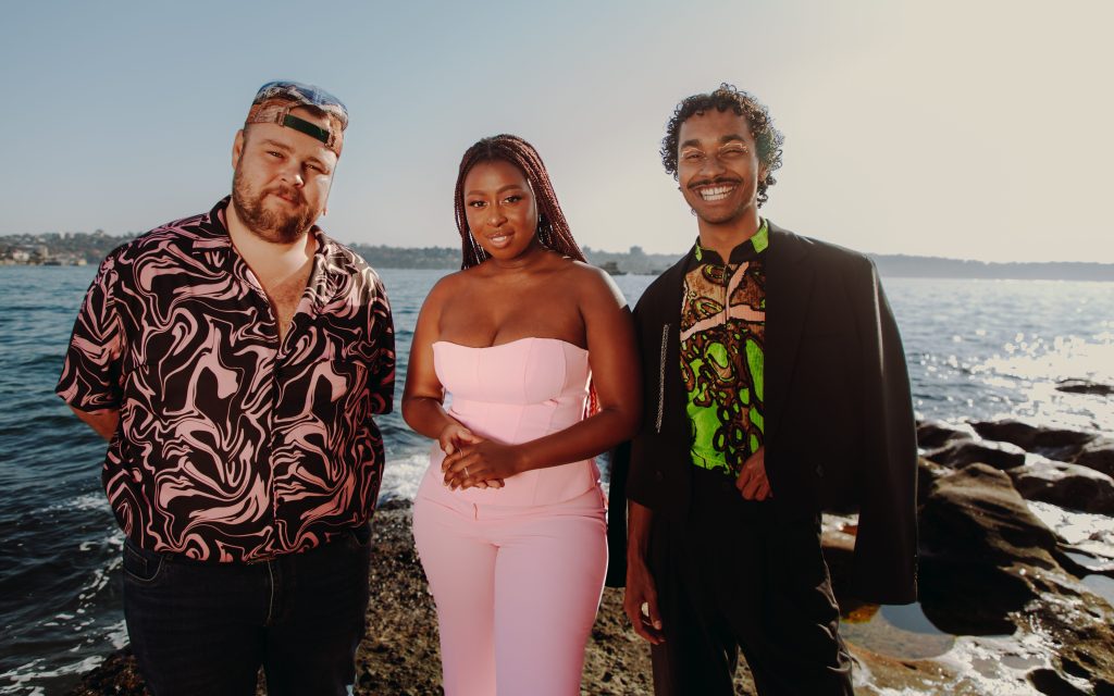 opportunities. Three people face the camera: a man in a patterned coloured shirt with a baseball cap, a woman in white shoulderless top and white pants and a man with a dark jacket.