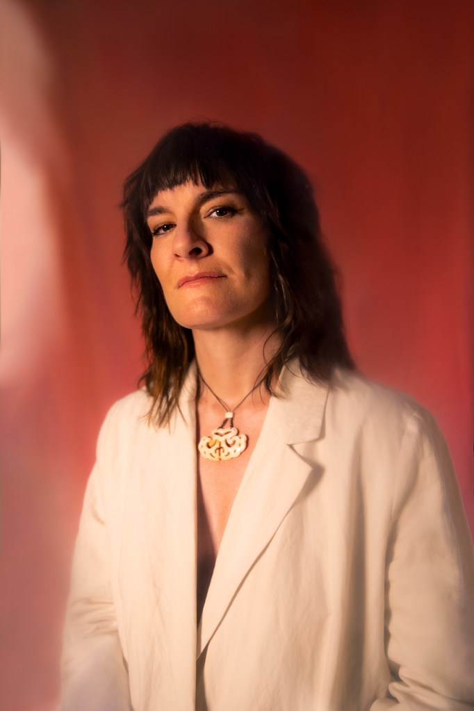 Woman with dark shoulder length hair and white jacket on reddish background