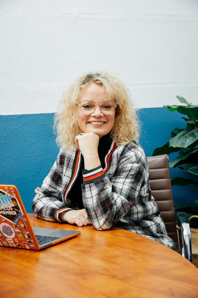 Woman with blonde wavy hair and glasses wearing black and white chequered jacket.