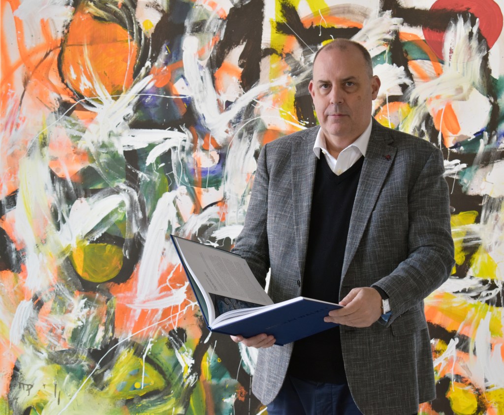 Man in black jumper and grey jacket holding open book in front of green orange white and grey artwork.