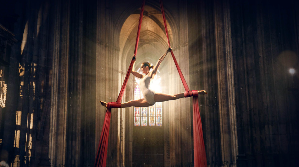 Woman in white doing the aerial splits on red silks against a stained glass window.