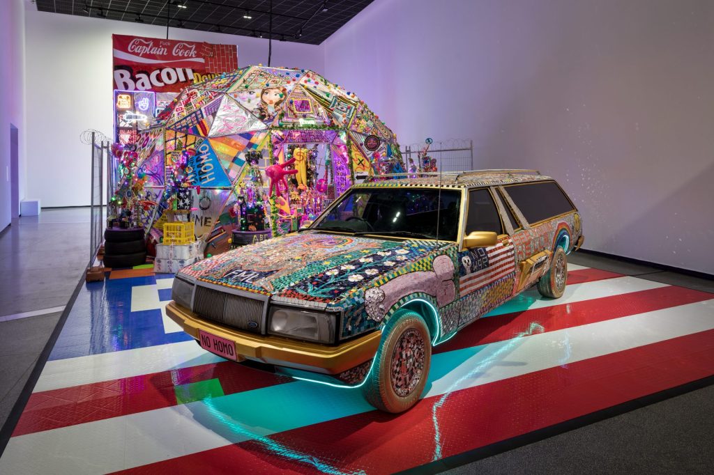 Multi-coloured car as artwork in museum on top of red and white striped floor.