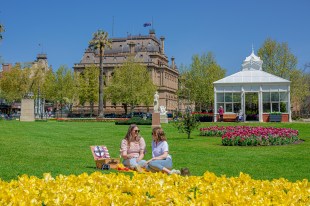 Two white women picnic on a green lawn, with bright yellow tulips blooming in the foreground. More tulips, and striking Victorian-era buildings, can be seen in the background.
