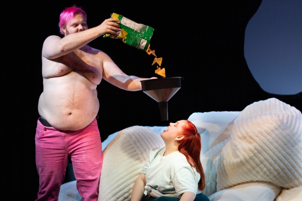 Fatness. A shirtless man with pink hair pours chips into a funnel for a woman with red hair and a white shirt to eat.