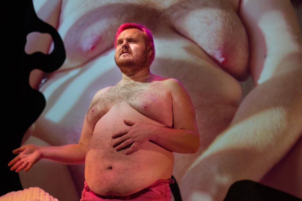 Fatness. A shirtless man with pink hair has one hand on his belly.