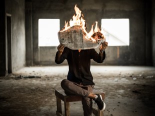 A man sits with one leg crossed over the other on a wooden chair in an empty warehouse. His face is obscured by the newspaper he is reading. The newspaper is on fire, suggesting he is reading this week's hottest arts news.