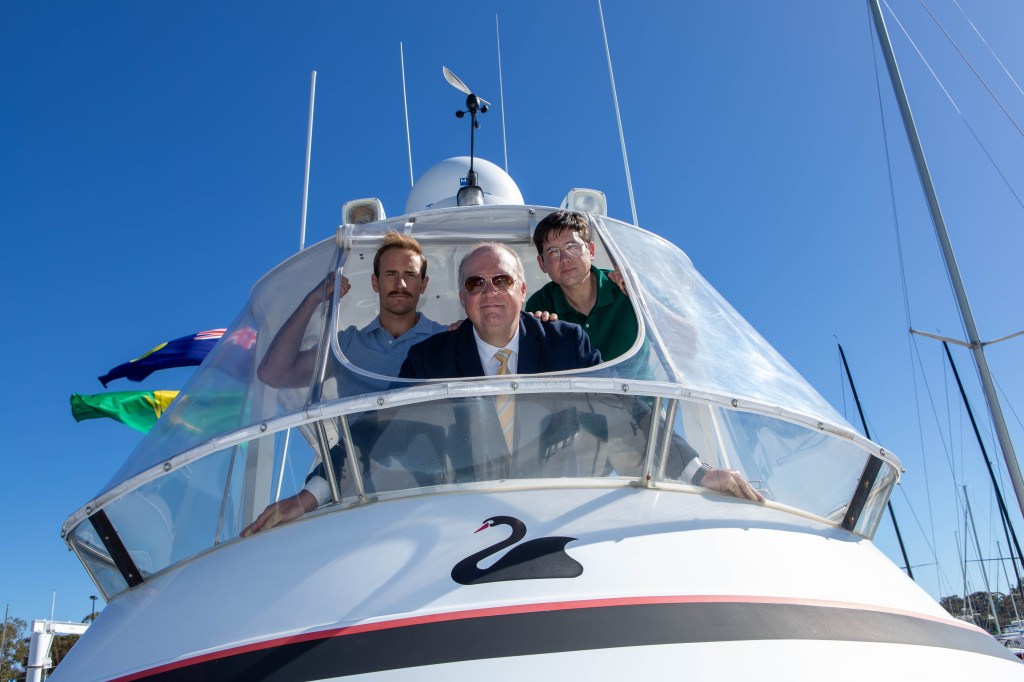 Three cast members of the production Taking Liberty are in a yacht and looking direct to camera.