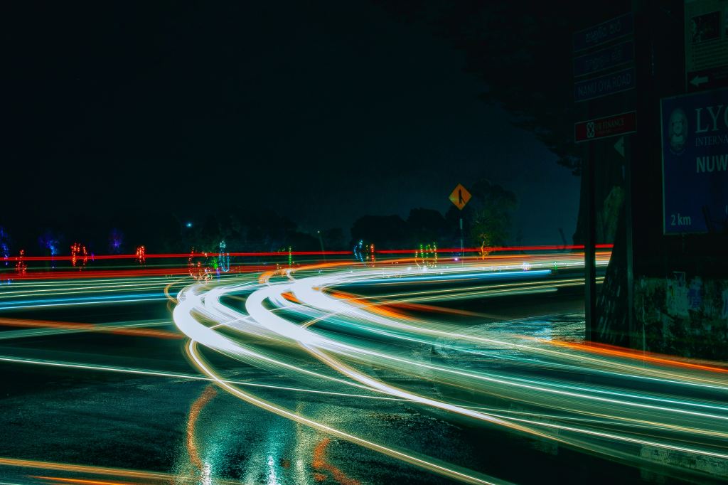 A long exposure photo of a wet road at night: car headlights and taillights are blurs of light reflected on the wet tarmac as well as floating between the road and the dark sky.