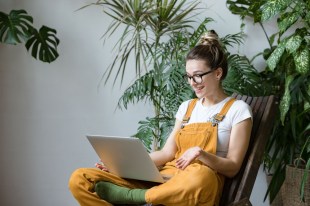 Arts news. Girl surrounded by plants reading from laptop and wearing yellow overalls.