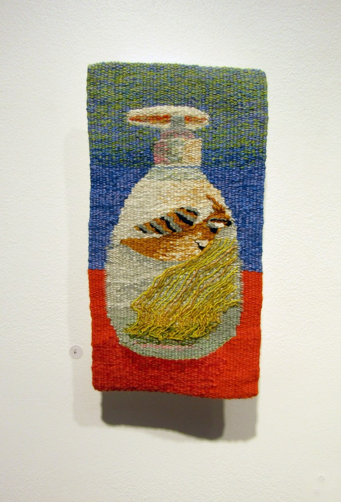 Short Shelf Life. Image is of green, blue and red background tapestry with a spray bottle and a nesting bird on it.