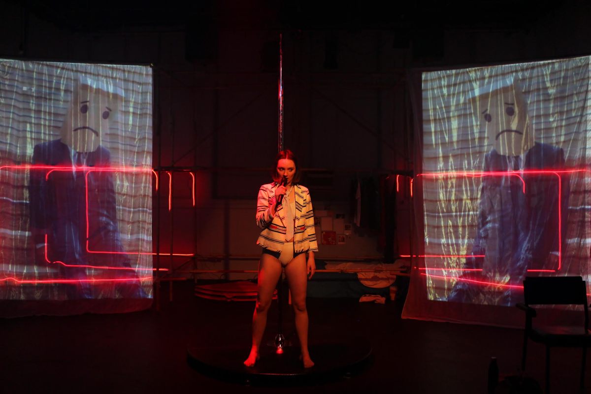 Gunasekera. Production still from ‘You're So Brave’. Photo: Rama Dolman. The image shows a female figure holding a microphone in the centre against a stage with red light and two projector panels on each side.