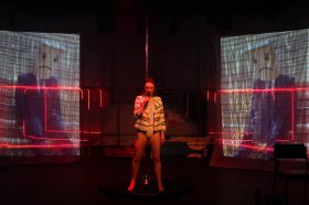 Gunasekera. Production still from ‘You're So Brave’. Photo: Rama Dolman. The image shows a female figure holding a microphone in the centre against a stage with red light and two projector panels on each side.