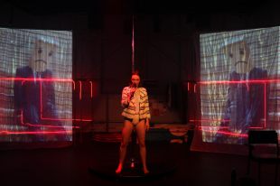 Gunasekera. Production still from ‘You're So Brave’. Photo: Rama Dolman. The image shows a female figure holding a microphone in the centre against a stage with red light and two projector panels on each side.