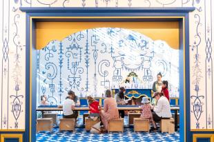 Children seated around a table with some adults, doing art activities with a blue and white wallpaper backround. They are at the Queensland Gallery of Modern Art Children's exhibition of 'Natalya Hughes: The Castle of Tarragindi. Art education for kids.