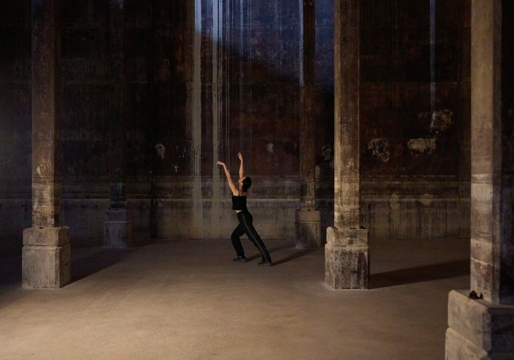 Angela Goh. Image is of a black clad dancer with arms upstretched at the back of a wide empty stage girt by colums.