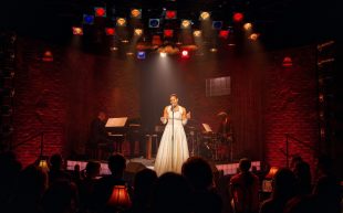 Lady Day. Image is a woman in a white dress backed by a trio singing into a mic in front of an audience.