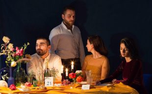 The Hotline. Three people sit at a table laden with food while a man stands behind them.