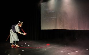 Love Letters. Image shows actor in 'A friendship of life and death' at Melbourne Fringe, bending character on empty stage wearing green costume with black sash and holding red string.