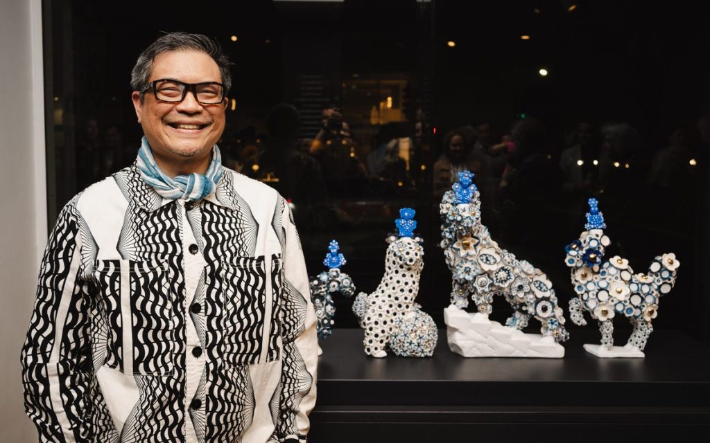 opportunities. Smiling man in white and black shirt with glasses standing in front of his mid sized sculptures.