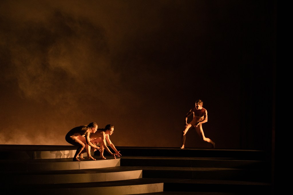Architect of the Invisible. Three dancers in beige costumes crouch on steps against an expansive brown backdrop.