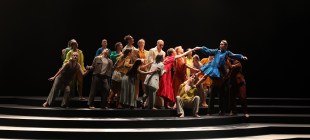 Architect of the Invisible. A large group of dancers in bright clothing reach across steps and lift dancer in blue dress.