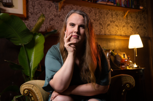 Trans. Woman with green top and long brown hair sits in armchair looking at camera with hand under chin. Behind her are a pot plant and a lamp.