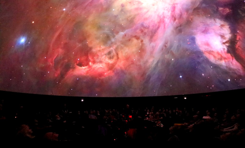 Angel Strings. Image is a cosmic view of swirling colours on a large screen on a dome, with a crowd seated below and looking up at the dome.