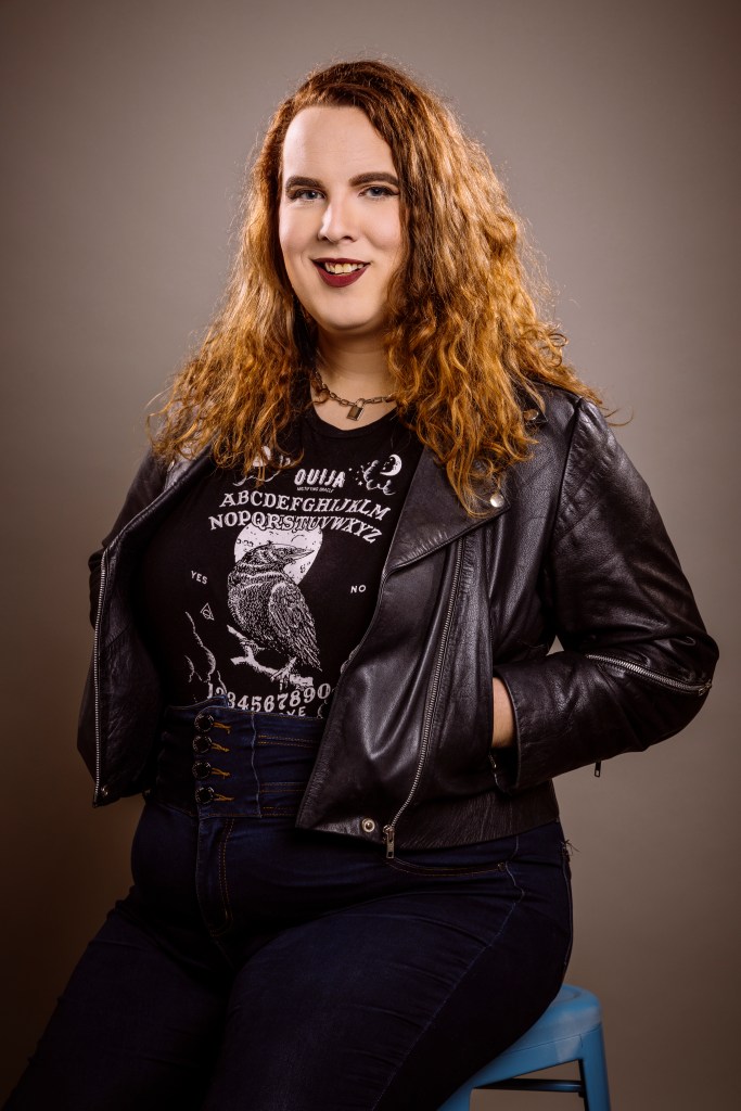 Trans. Woman sitting on stool wearing black leather jacket and with hands in pockets.