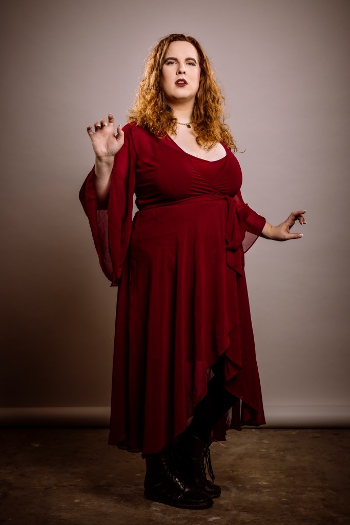 Trans. Woman in flowing dark red dress standing with raised hands.