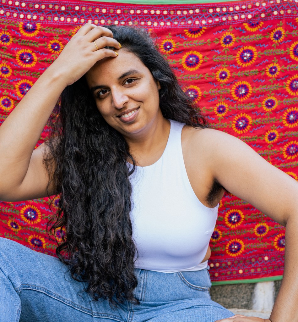 path. Image is of a woman of Asian appearance with long dark wavy hair sitting in front of a red patterned cloth backdrop, wearing jeans and a white sleeveless top. She has one hand on her head and is smiling.
