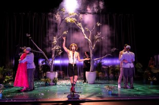 Twelfth Night. A dark spotlit stage features small trees in large pots, a couple of actors cuddle on. either side of the stage and a person centre stage with arm raised, wearing a white waistcoat, braces and black shorts.