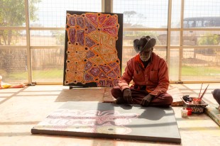 Uplands. Aboriginal man wearing hat and sitting on ground in an art centre with painting around him