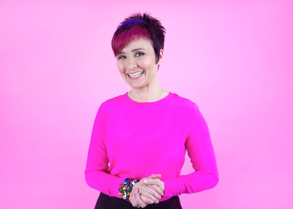 Cal Wilson. Image of woman with short red and black hair, wearing pink jumper and clasping hands.