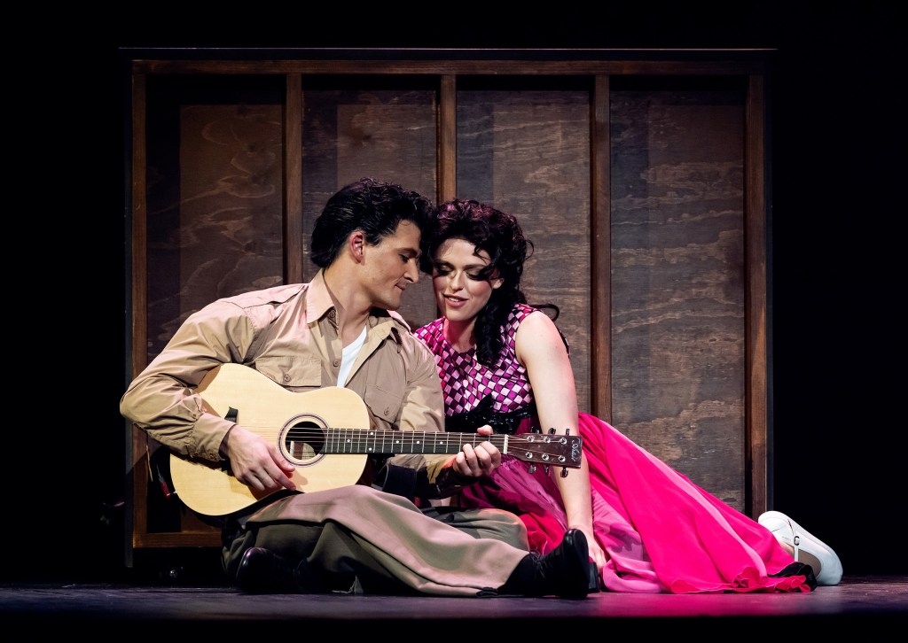 Elvis. Man in army fatigues playing guitar and talking to girl in pink dress, sitting next to him.