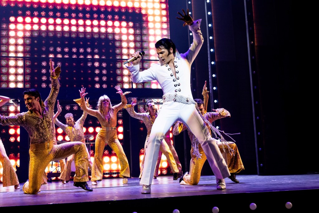 Elvis. Man in white jumpsuit singing into a mic with dancers in gold lame behind. 