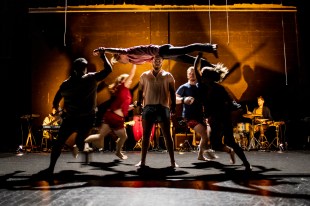 Australian Circus Summit. Image is of a dark stage with some spot lighting and a band along the back wall. Centre stage is a man standing with four others running around him and one other person flat out and held aloft.