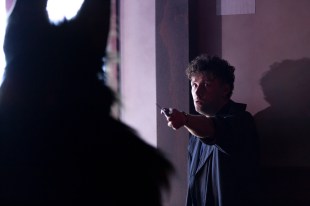 Hour of the Wolf. Image shows the back of a silhouetted wolf figure and a man with his back to the wall brandishing a sharp object at it in fear.