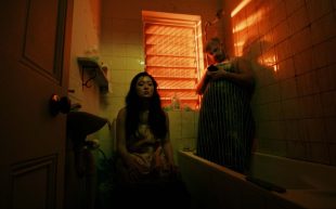 Sweet Juices. Image is of a dark bathroom with orange light coming through a slatted window. A woman of Asian appearance and long dark hair sits on the toilet looking despondent, while a bearded man in a striped apron stands in the bath looking at his phone.