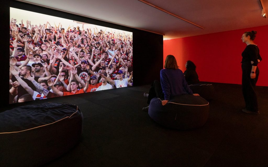Collective Knowledge: image is a screen depicting many people with arms outstretched three people with a red wall behind them looking at the screen.