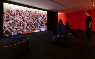Collective Knowledge: image is a screen depicting many people with arms outstretched three people with a red wall behind them looking at the screen.