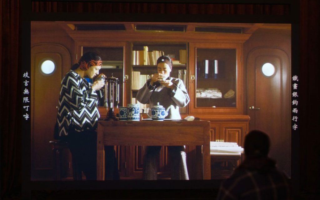 Image is of a person watching a screen on which two women in China are drinking tea at a table in front of a glass and wood cabinet that contains artefacts and books.