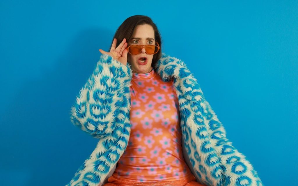 Big Dyke Energy. Image is woman in orange and blue dress with furry blue and white coat, orange shades on the end of her nose and mouth agape in a shocked expression.