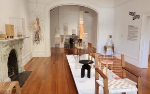 Design Fringe. Image is white gallery space and wooden floor and empty fireplace, with tables and shelves covered with art objects and pieces of furniture design.