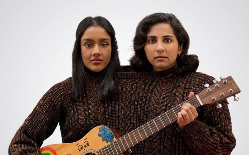 The Coconuts. Image is two women with brown skin wearing the same big brown jumper and holding a guitar.
