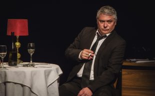 Ink. Man in suit and tie, sitting on stage next to a fancy dining table, smoking a cigarette.