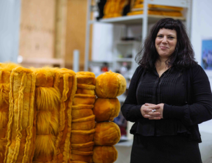 Education for arts teachers. image shows woman in black with hands held in front of her and shoulder length dark hair smiling and standing next to yellow soft sculpture of interwoven bales and strands of faux fur.