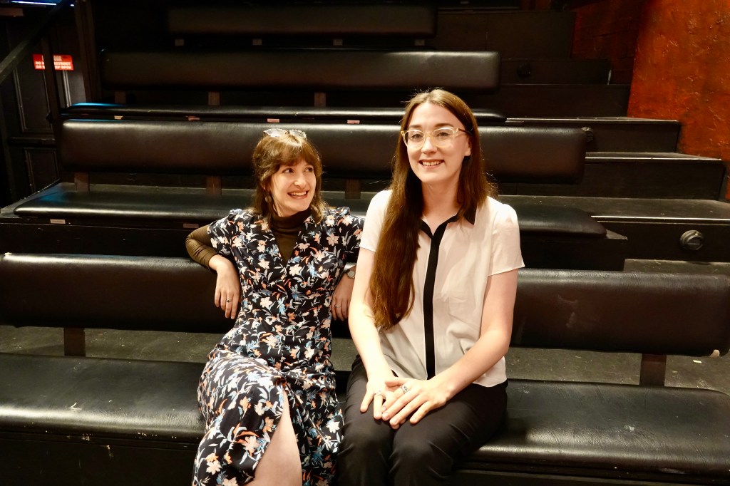 Old Fitz. Image is rows of bench theatre seating, with two young women sitting on the front row, one in a patterned dress and one in a white shirt and dark skirt.