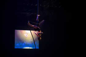 Circus. Image is a man hanging upside down from a rope against a black background.