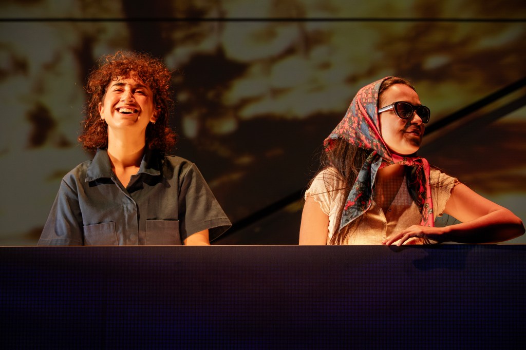 Girls in Boys' Cars. Image is two young women, one with shoulder length hair grinning and wearing a blue short sleeved shirt and the other looking off to the right in sunglasses and headscarf. They are seated behind a block, representing a car.