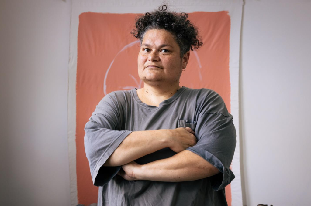 Pacific art. Image is of a person with dark curly short hair, folded arms and grey shirt looking askance at the camera and standing in front of an ochre coloured wall hanging.